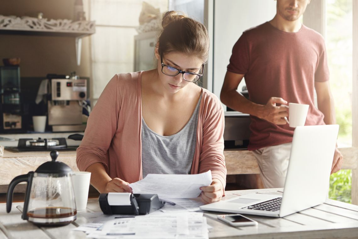 Family budget and finances. Young woman doing accounts together with her husband at home, planning new purchase. Serious female in glasses holding piece of paper and making necessary calculations